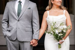 Close-up of bride and groom holding hands, groom in gray suit with green tie, bride holding wildflower bouquet.