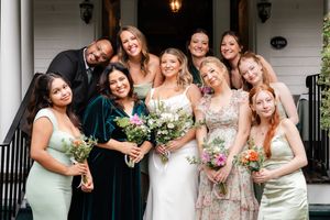 Bride surrounded by bridesmaids in sage green and floral dresses holding colorful bouquets outside white venue.