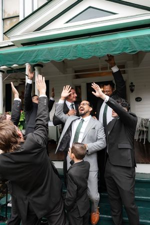 Groom in gray suit celebrates with groomsmen in black suits raising hands under green awning.