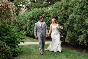 Bride and groom walking hand in hand through lush garden path, bride in fitted white gown with bouquet.