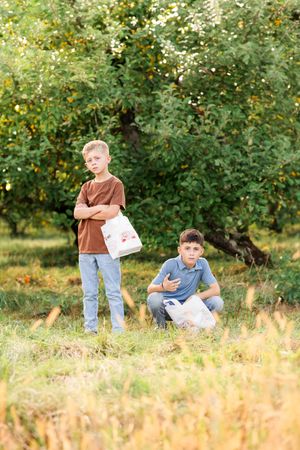 Two boys holding bags standing in green apple orchard during summer harvest.