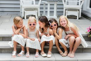 Five young girls in matching outfits and pink sunglasses sitting together on stone steps, laughing and smiling.