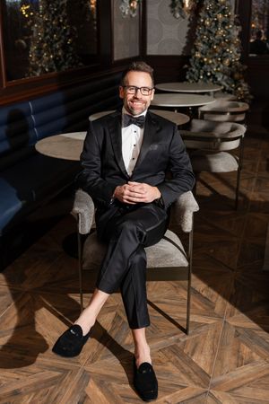 Dapper groom in black tuxedo with bow tie sitting confidently in a chair at an elegant venue with Christmas trees.