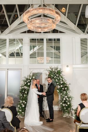 Bride and groom exchanging vows under a grand crystal chandelier at a bright, modern wedding ceremony venue.