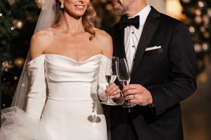 Newlywed couple in wedding attire toasting with champagne glasses near a festive Christmas tree backdrop.