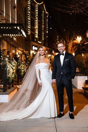 Bride in off-shoulder gown with flowing veil and groom in tuxedo posing on a city street with holiday lights in downtown Saratoga Springs.