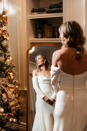 Bride in elegant off-shoulder white gown admiring her reflection in a gold-framed mirror beside a decorated Christmas tree.