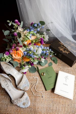 Wedding flat lay featuring a colorful bridal bouquet, embellished heels, vow books, and gold rings on woven mat.