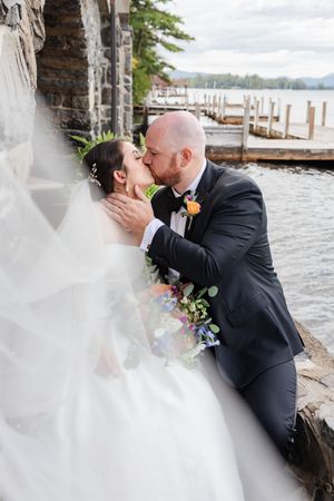 A bride and groom share a kiss against a stone wall with a lake and dock in the background.