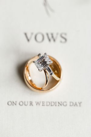 A princess-cut diamond engagement ring rests inside a gold wedding band on a vows booklet.