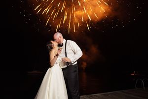 A bride and groom share a romantic kiss on a dock beneath a stunning fireworks display at night.