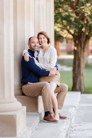 A smiling couple sits together against a stone column outdoors, embracing warmly in soft natural light.