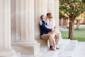 A couple shares a tender kiss while sitting together between elegant stone columns in a sunlit outdoor setting.