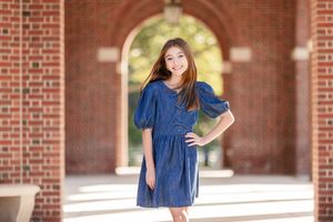A young girl in a blue denim dress poses confidently under a brick archway in warm sunlight.
