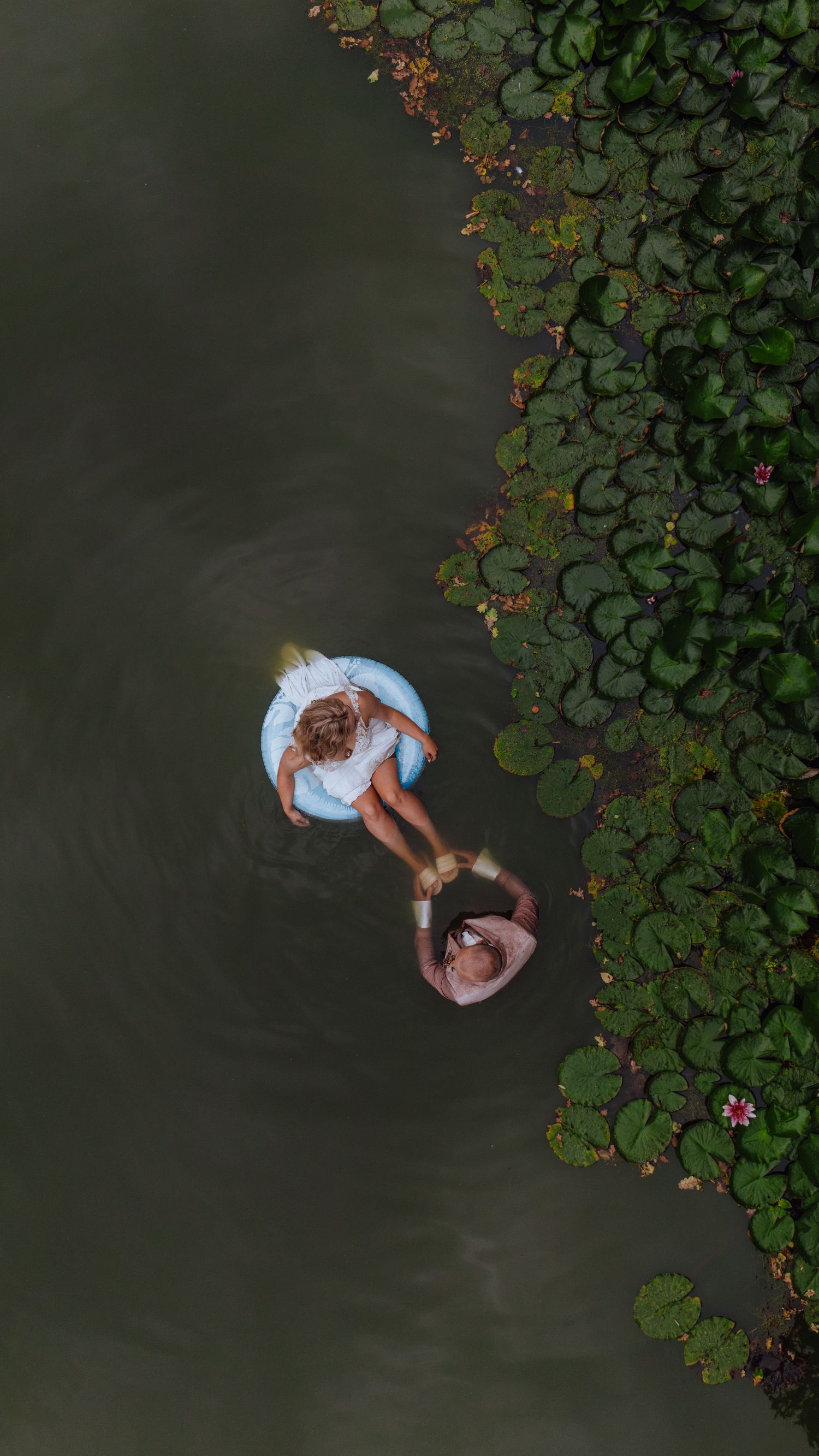 Luchtfoto van twee mensen die aan de rand van donker water zitten, naast waterlelies en gebladerte.