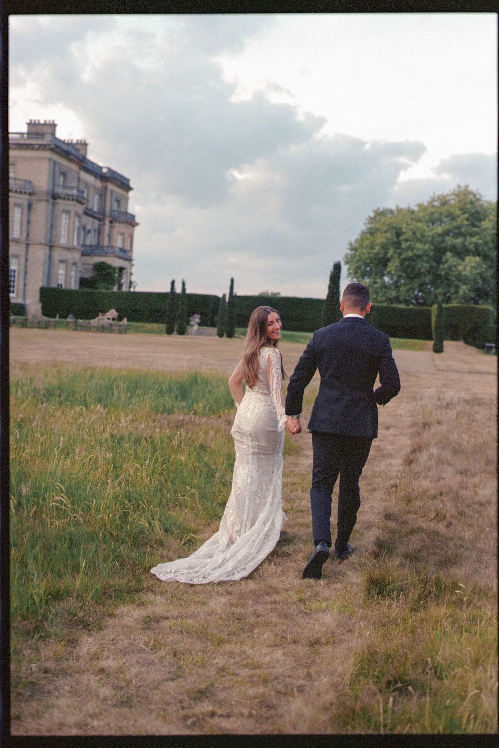 A couple walks hand in hand on a dirt path near a grand estate, with the bride wearing a flowing lace wedding gown.