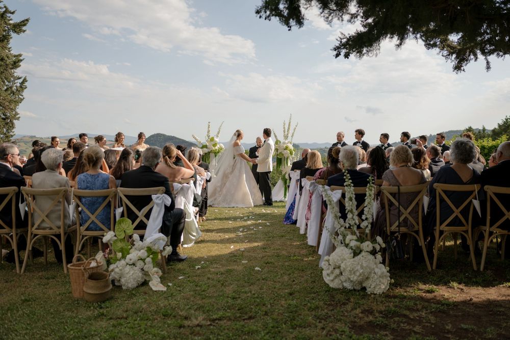 Outdoor wedding ceremony with guests seated on wooden chairs along a grass aisle, bride and groom at the altar with scenic views.