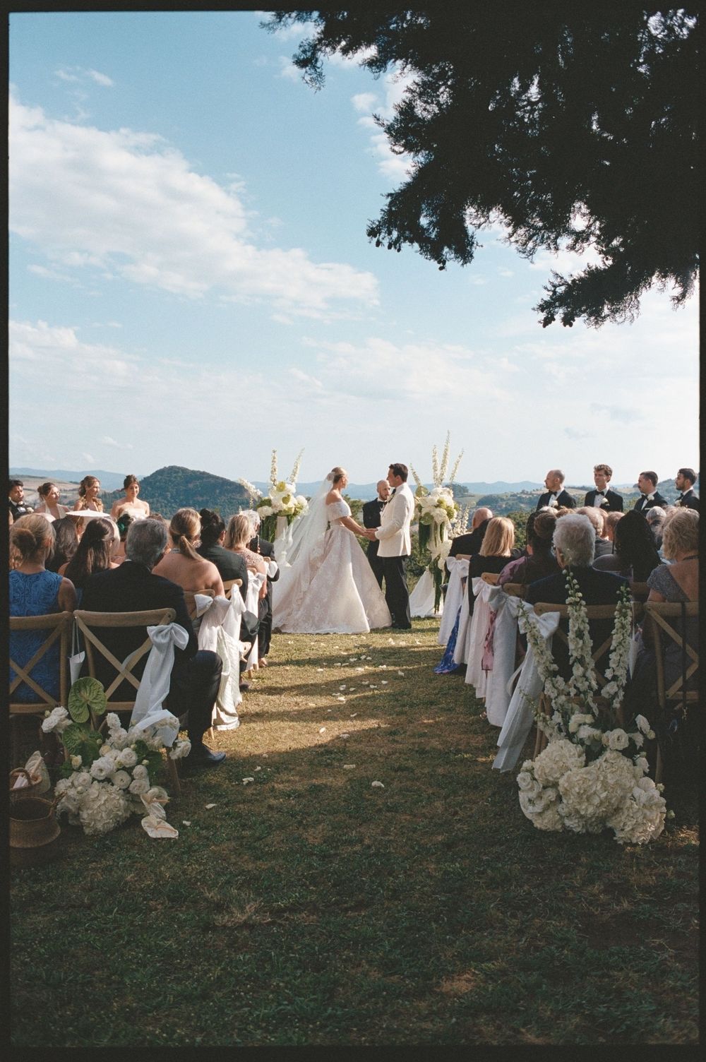 Outdoor wedding ceremony on a hillside with bride and groom at the altar, white floral aisle decor, and mountain views.