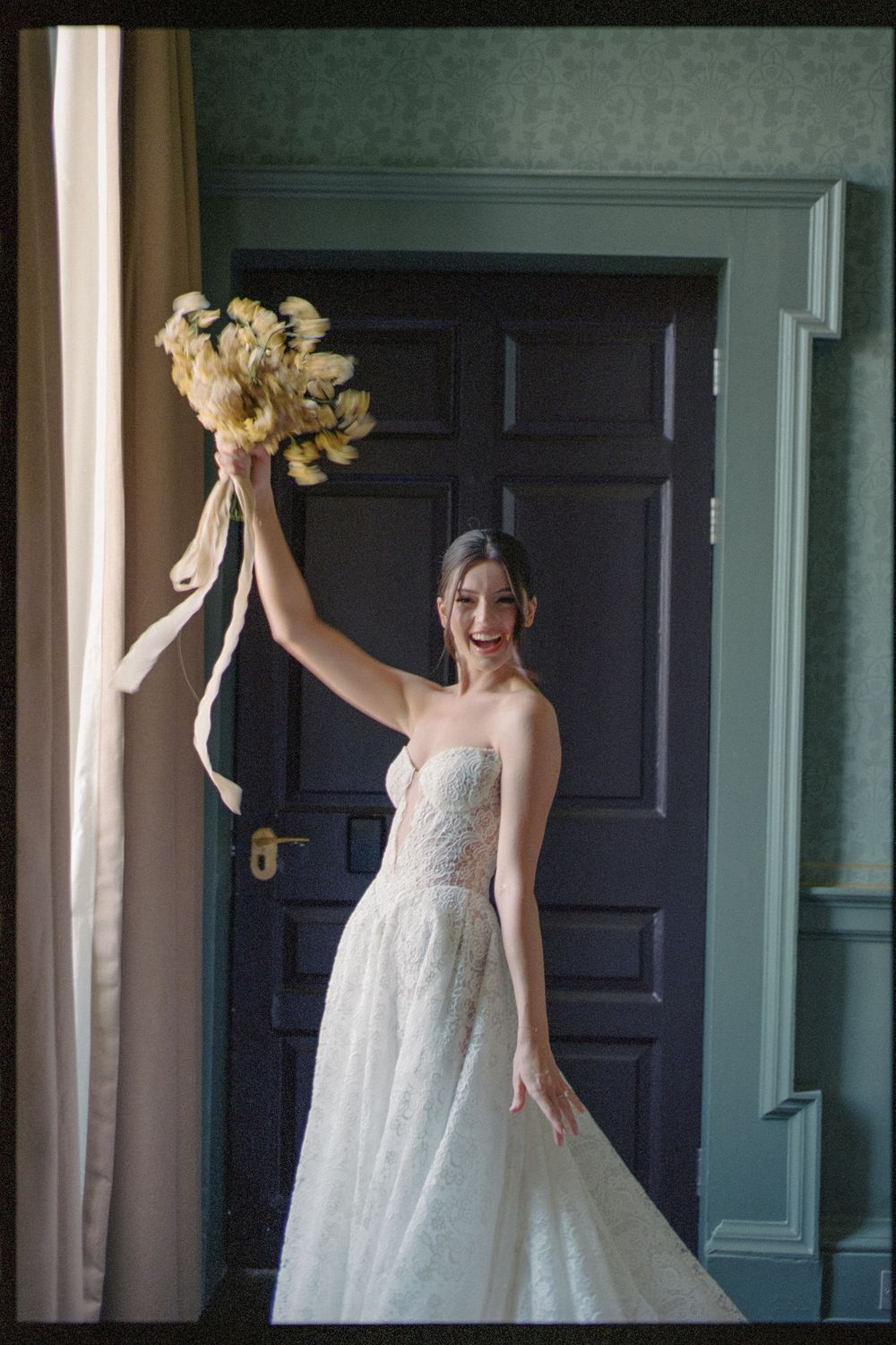 A bride in a flowing white wedding gown stands joyfully against a dark door in a mint-green room.