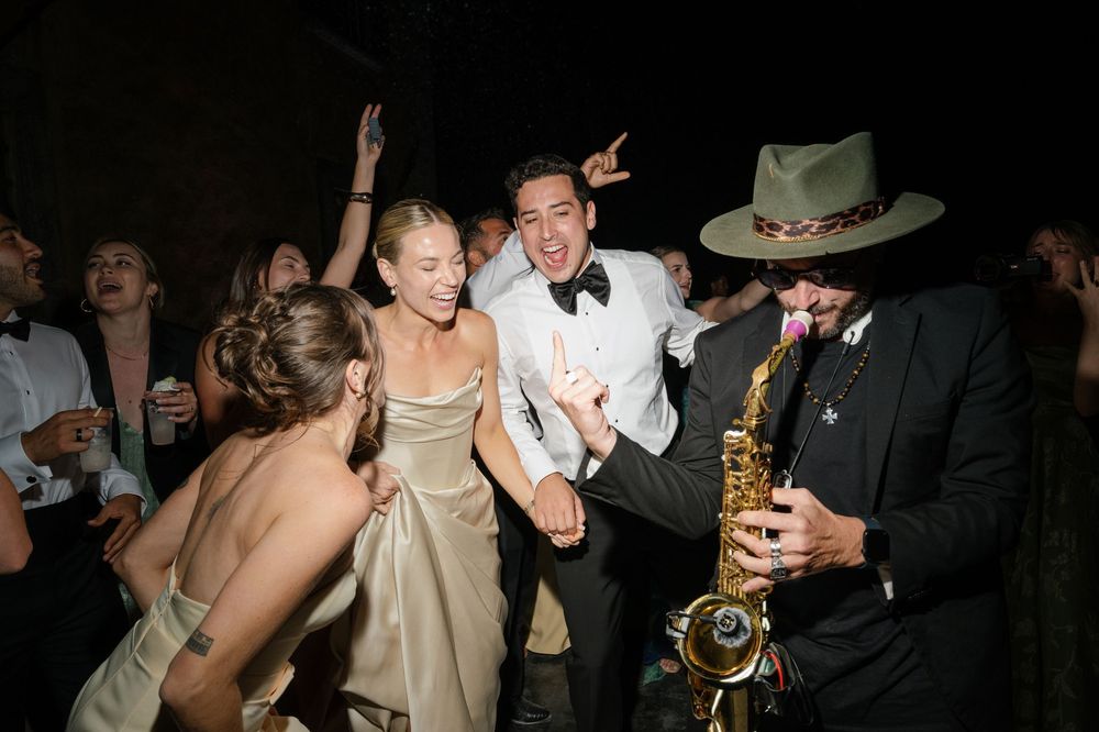 Wedding couple dancing joyfully with guests and a saxophonist at a lively evening reception party.