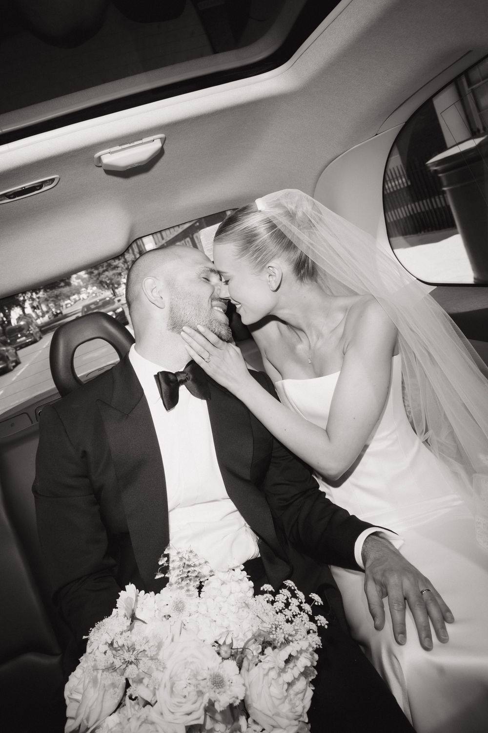 Black and white photo of a bride and groom sharing a romantic moment in a limousine on their wedding day.