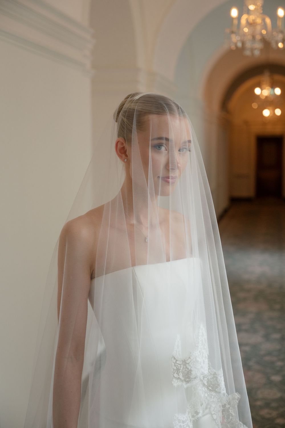 Bride in white strapless gown wearing a sheer veil over her face, standing in an elegant hallway with chandelier.