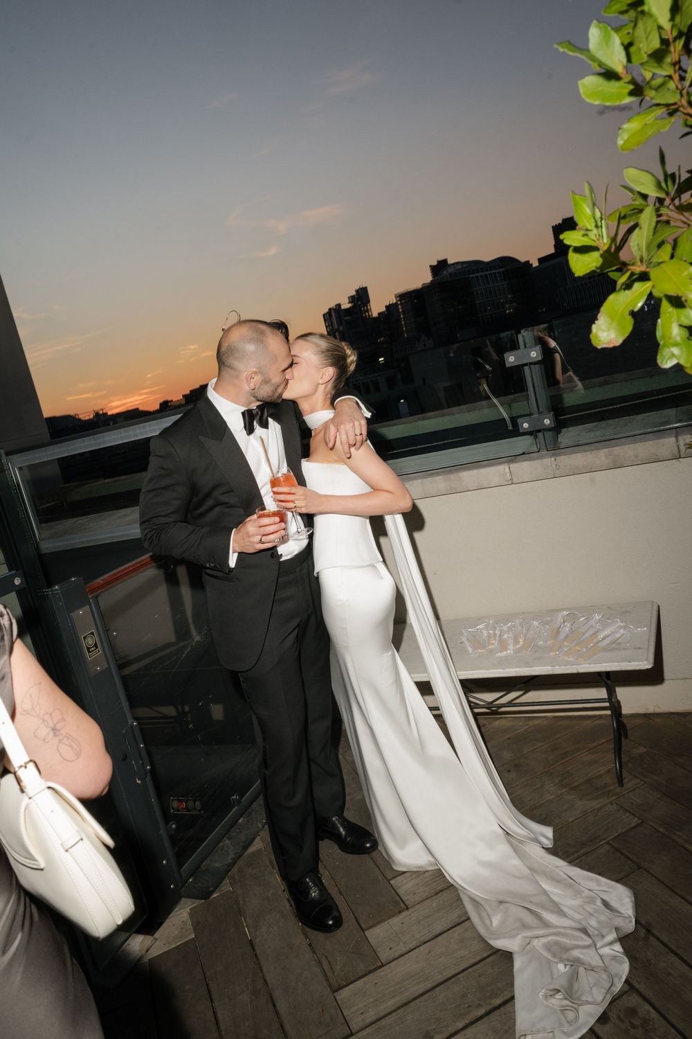 Bride and groom share a romantic kiss on a rooftop terrace at sunset, bride wearing elegant white gown with long train.