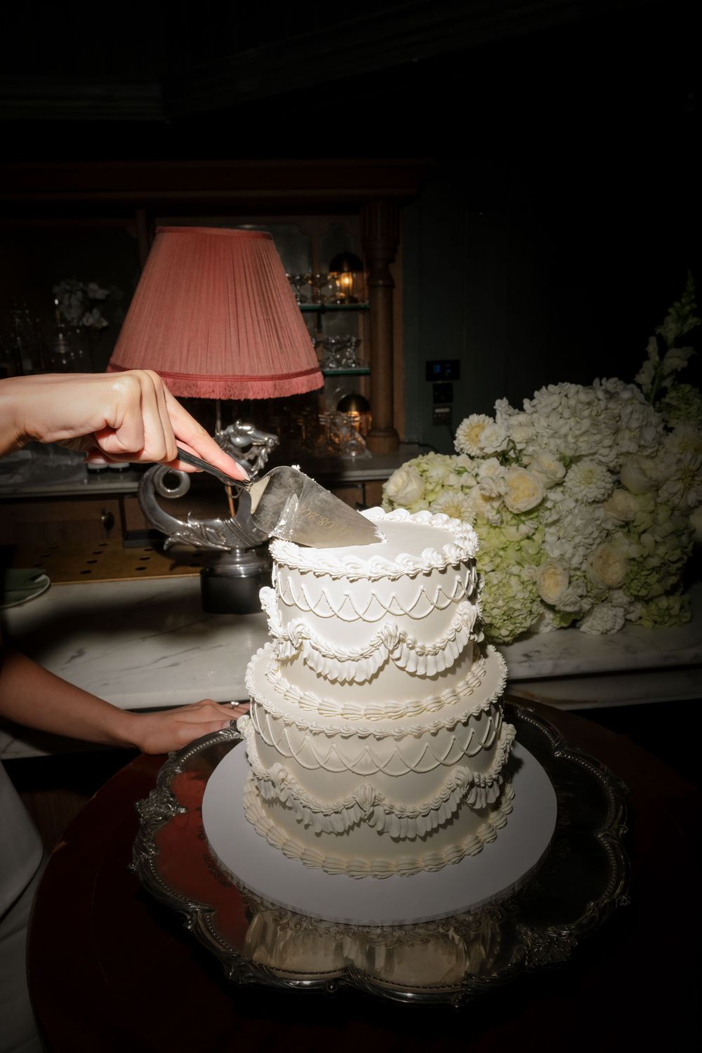 Hands cutting a white three-tier wedding cake on silver platter with floral arrangement and pink lamp in background.