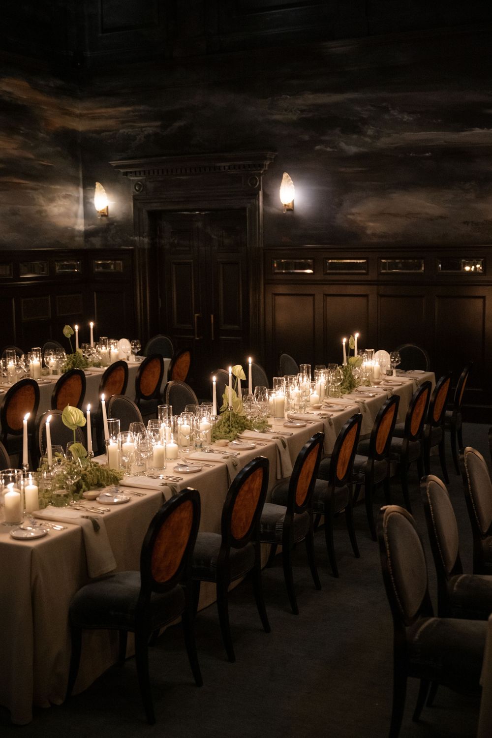 An elegant candlelit dining table set in a dark room with white linens and ornate chairs creates a romantic atmosphere.