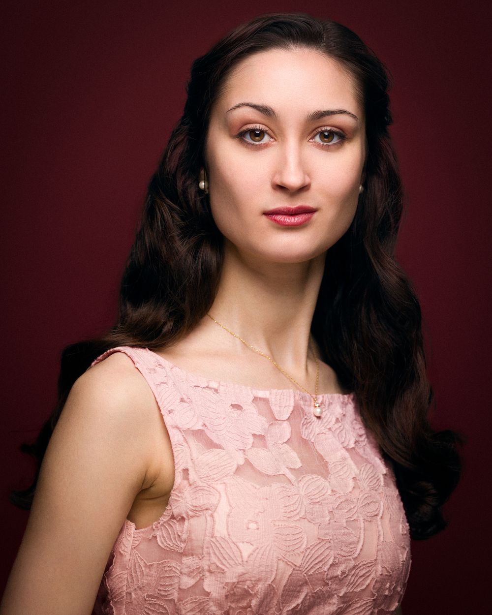 A woman with warm brown eyes and softly waved dark hair wears a pink lace dress and pearl jewelry, framed by a dramatic burgundy background.