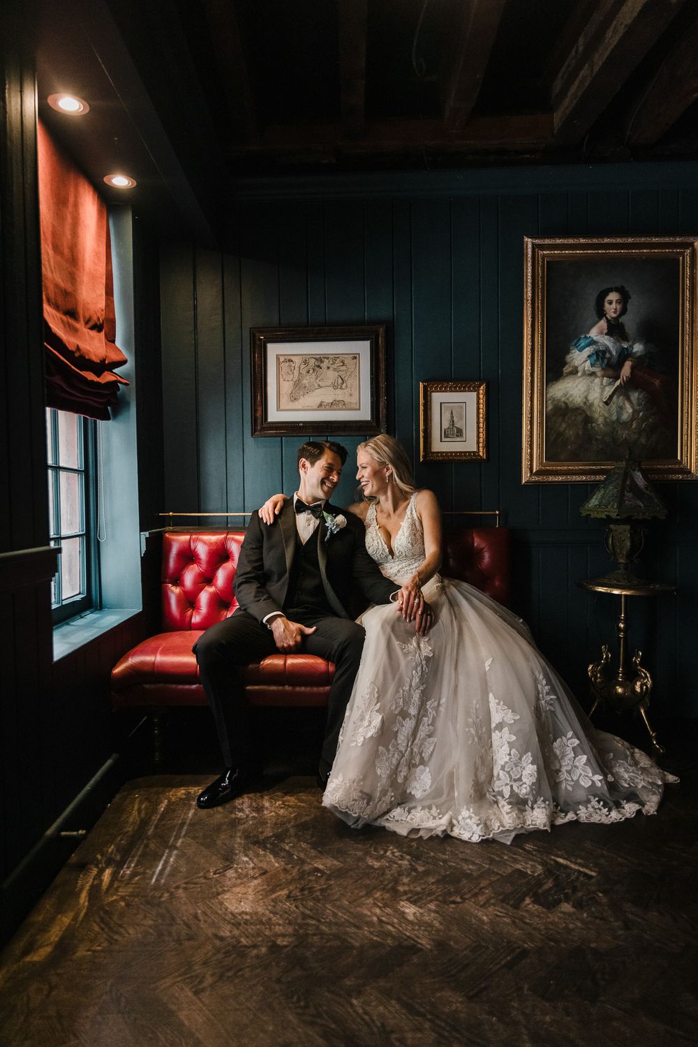 A bride and groom sit on a bright red love seat. They are lit softly with window light.