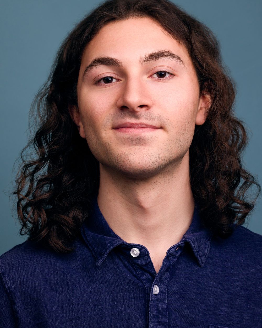 Young man with long curly hair, wearing a blue shirt, natural smile against a teal-blue backdrop.
