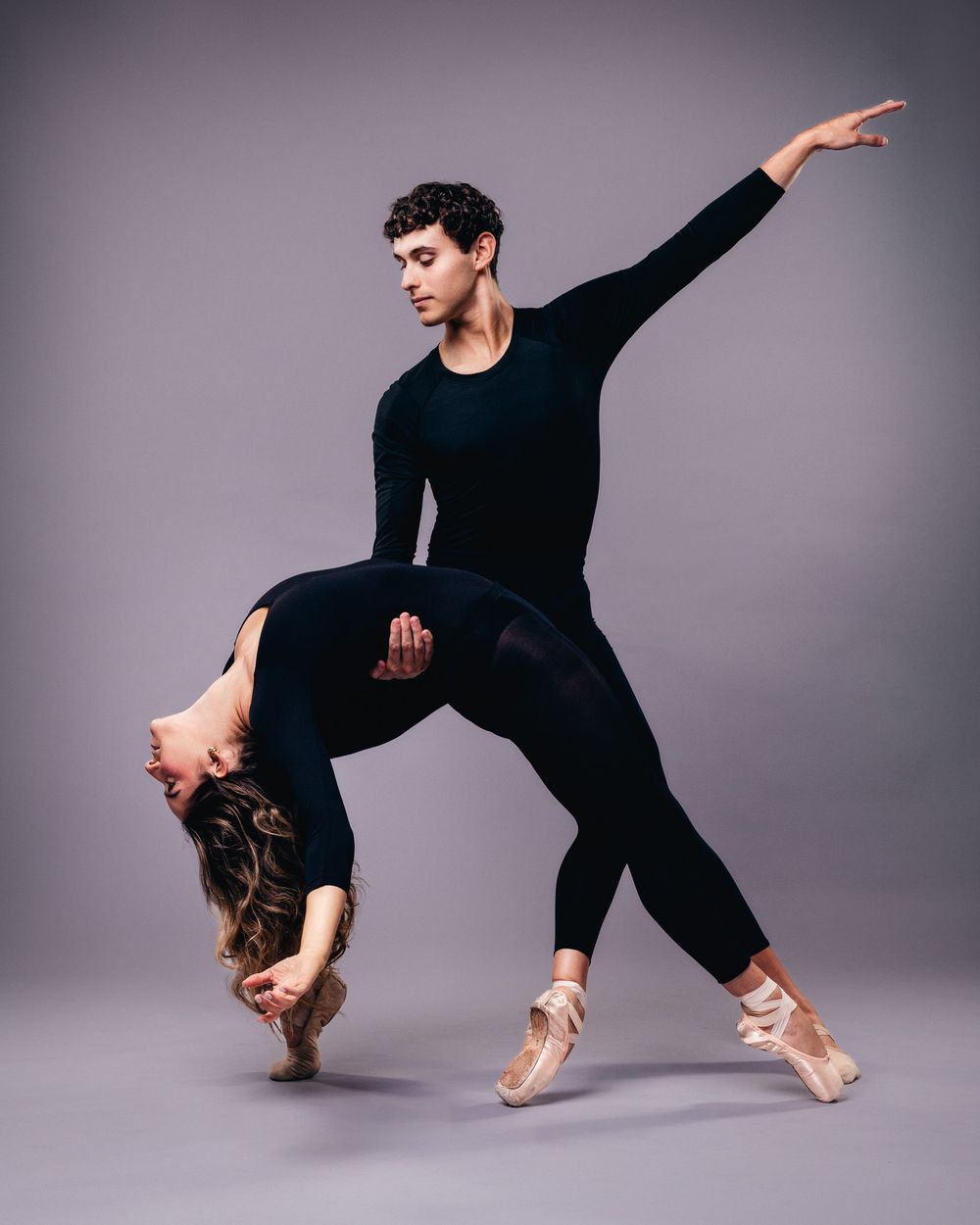Two ballet dancers in a dramatic pose, the woman arched backward midair as the man supports her, both in black against a gray backdrop.