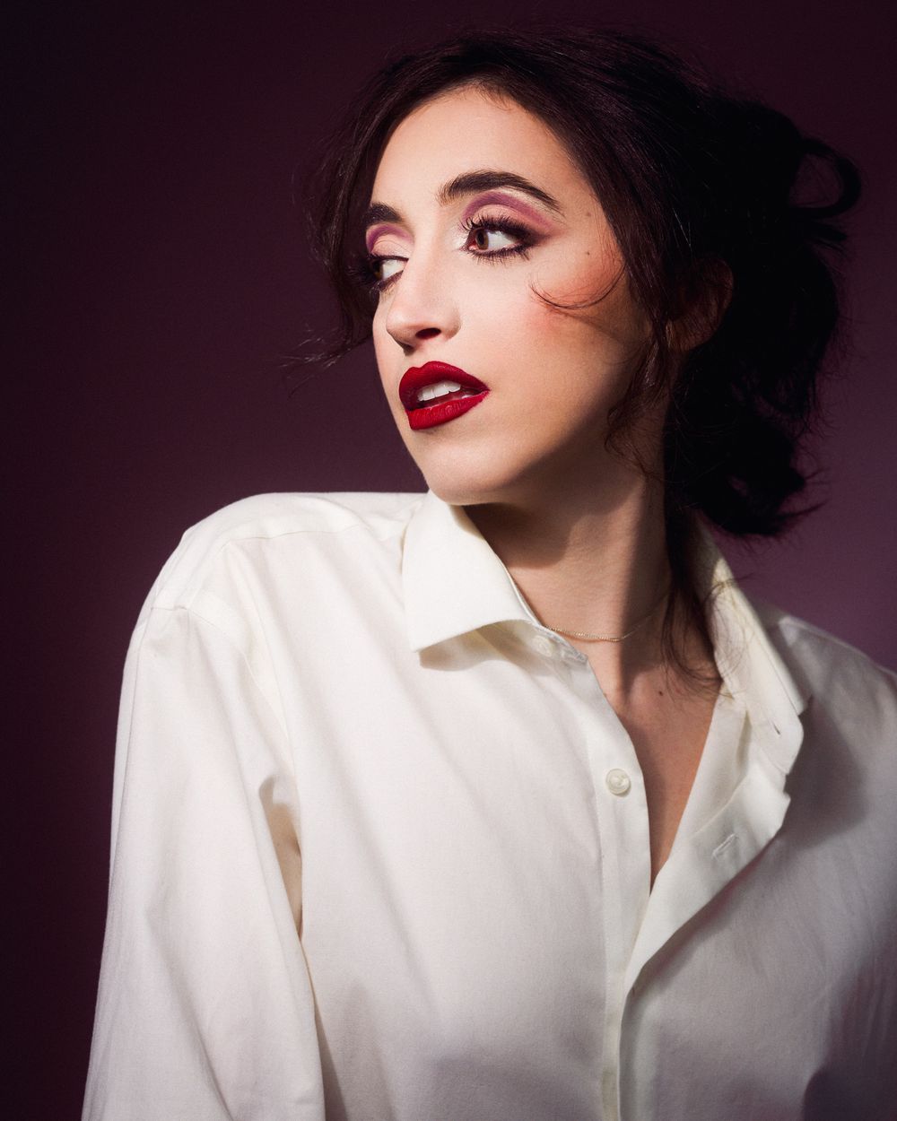 Woman in oversized white shirt, bold lipstick, styled with vintage flair against a plum-toned background.