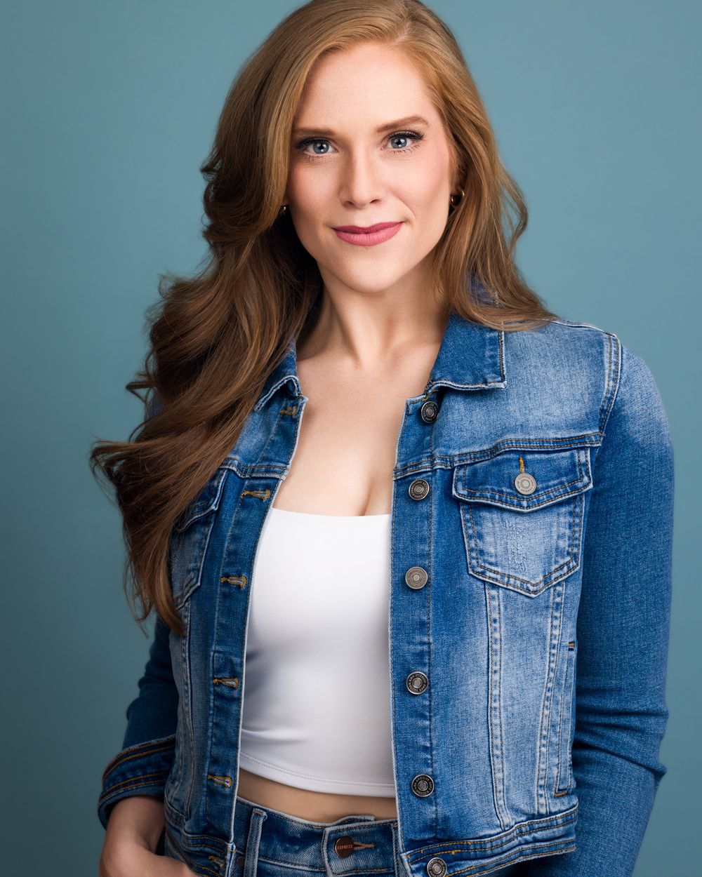 Confident woman with long auburn hair in a denim jacket and white crop top, smiling slightly against a soft blue background.