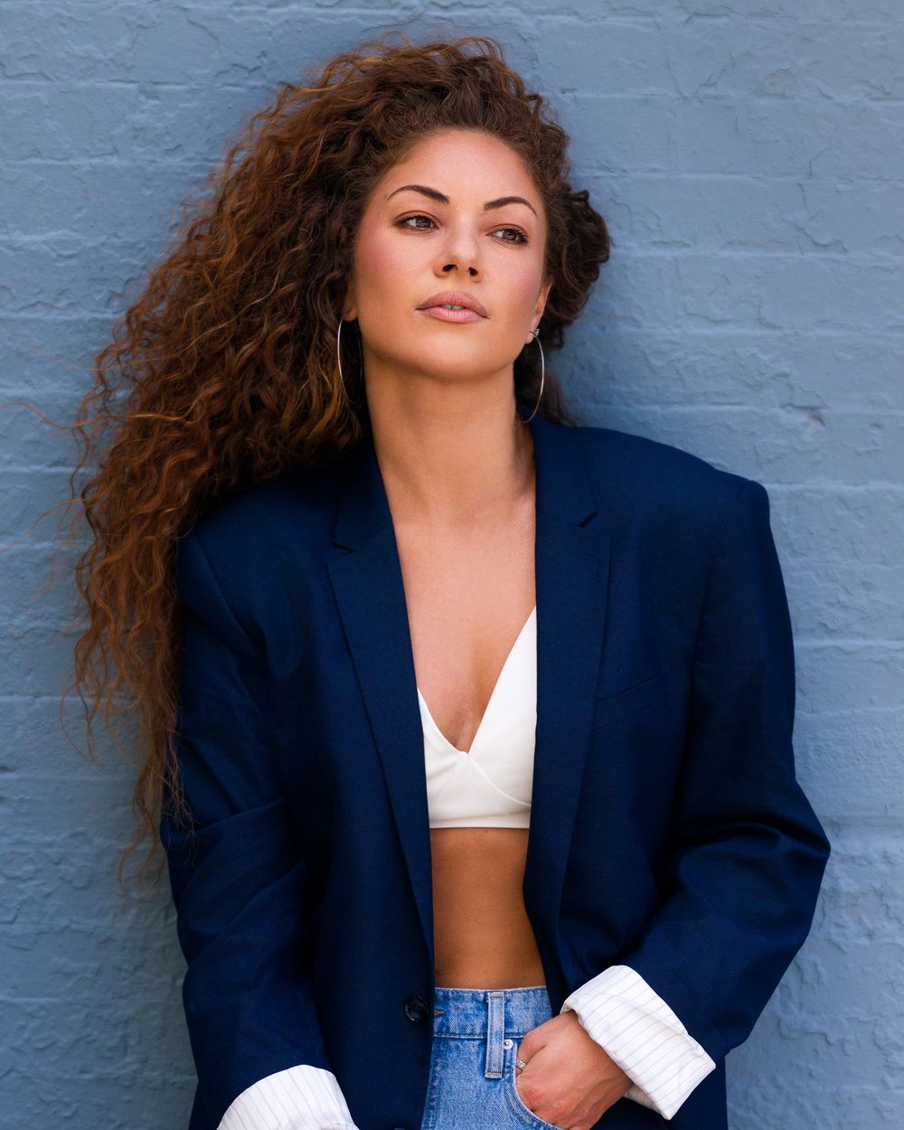 Woman with voluminous curls in a navy blazer and crop top, strong pose against a slate blue wall.