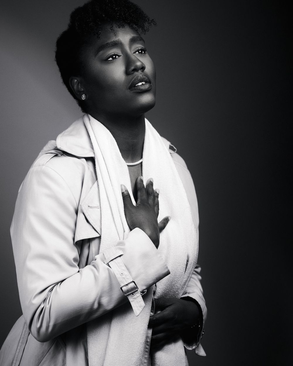 Dramatic photo of a woman in a white trench coat softly clutching her chest and staring longingly into the distance.