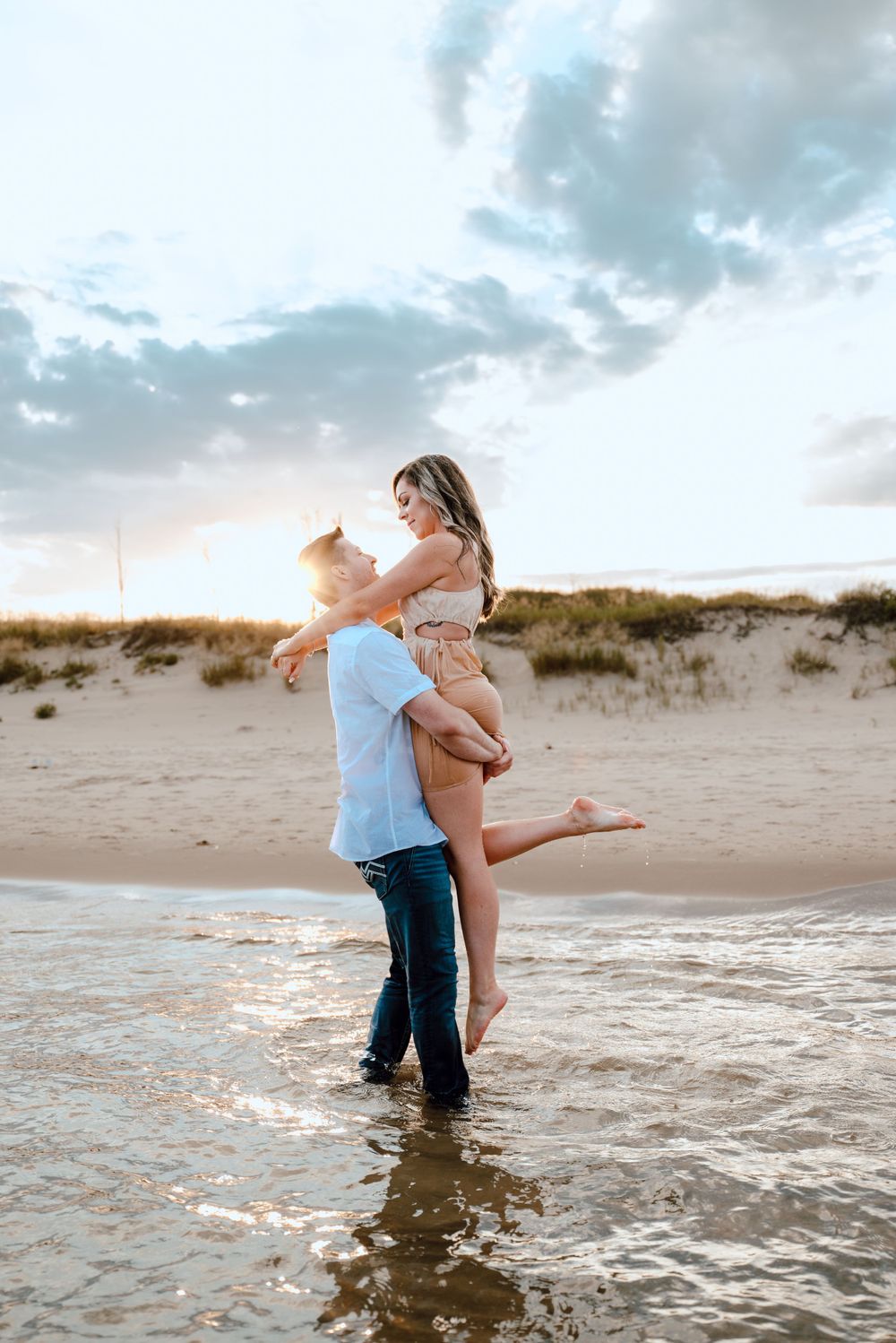 A romantic couple embraces in shallow ocean water at sunset with sandy beach and cloudy sky in background.
