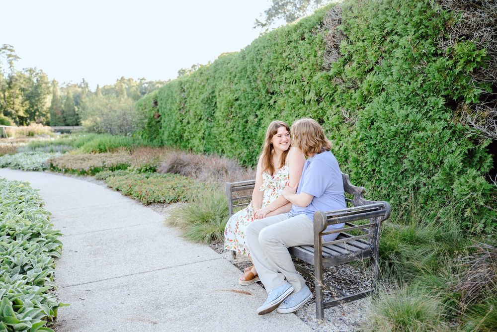 Two people sit together on a wooden bench along a winding garden path next to tall hedge walls in warm sunlight.