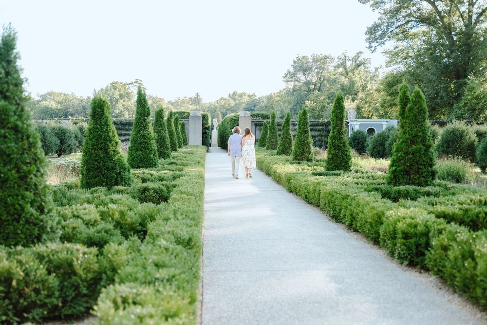 A winding garden path lined with manicured cypress trees and hedges leads through a tranquil formal garden.