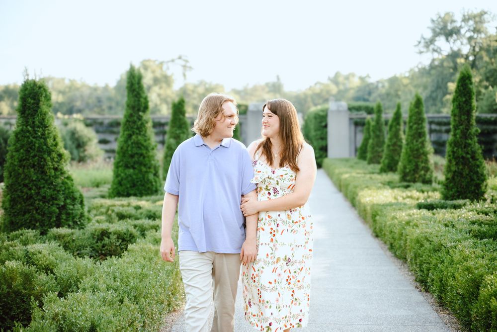 Couple strolls hand in hand on garden path lined with manicured evergreen trees and blooming flowers.