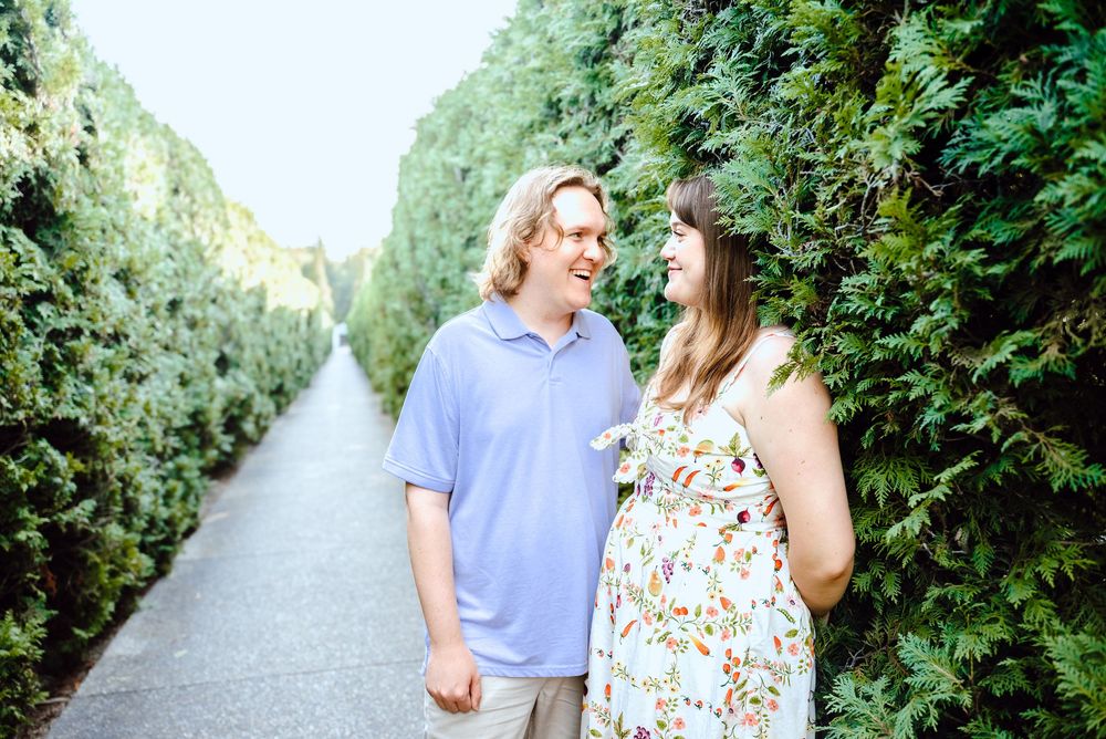 A couple walks along a scenic garden path surrounded by lush green trees and hedges.