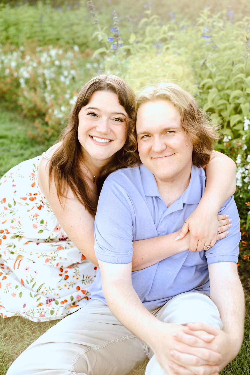 A couple sits together outdoors in a garden with blue flowers and greenery during a sunny summer day.