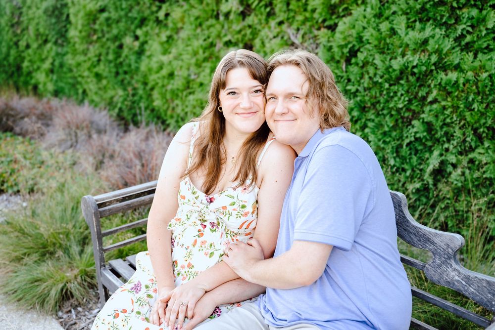 Two people embrace and smile warmly while sitting together outdoors surrounded by lush green foliage.