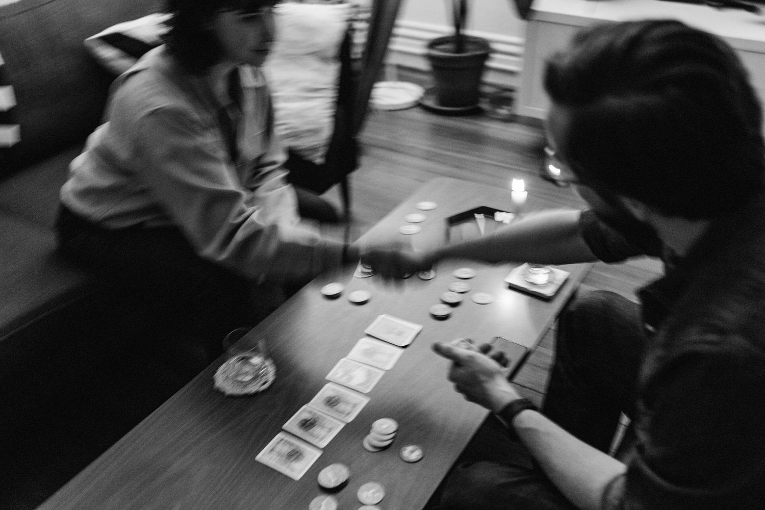 A black and white photo of a New England couple shaking hands over a table with a board game out on it. The photo has motion blur.