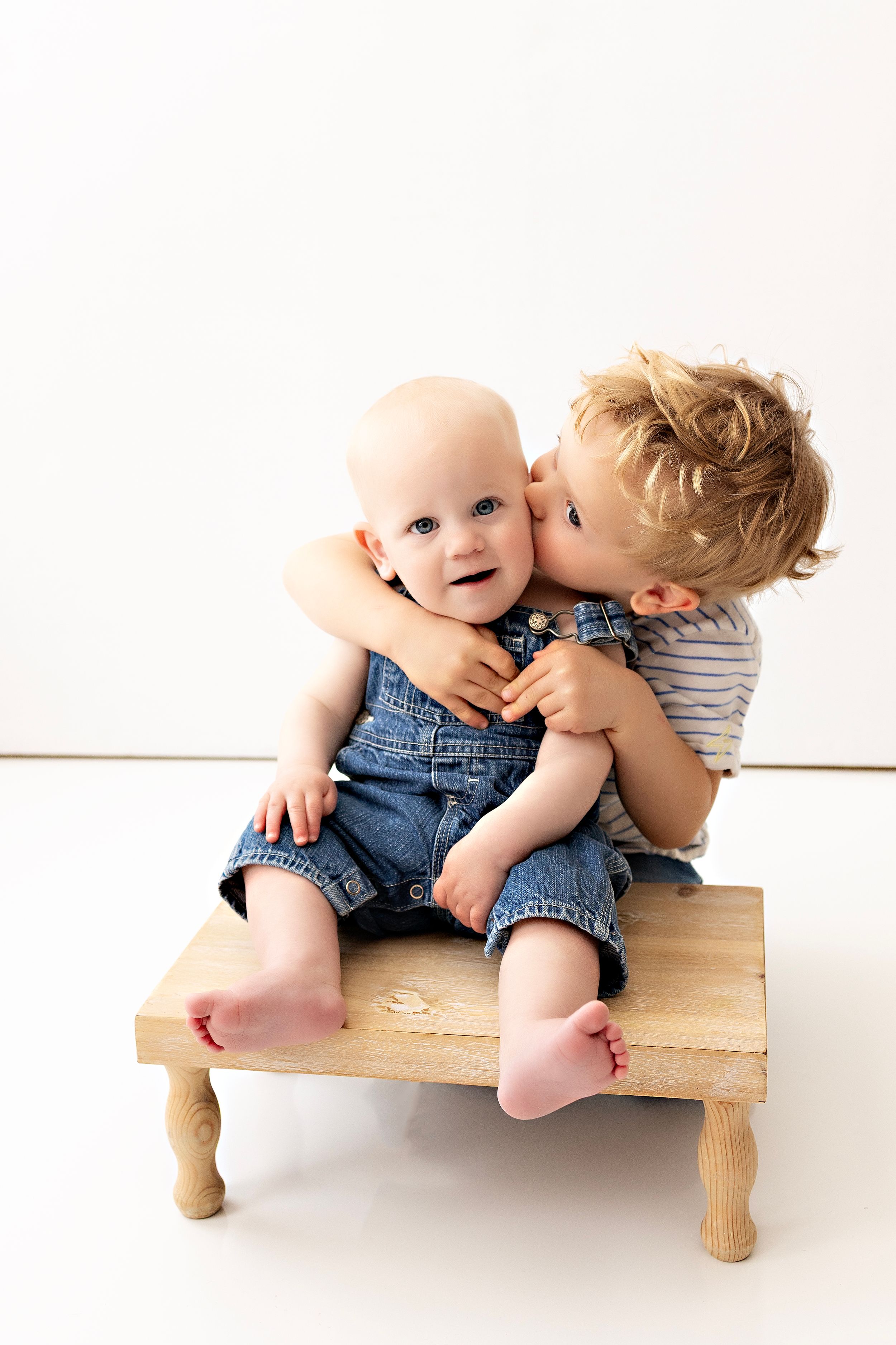 Two toddlers share a sweet moment sitting on a small wooden stool against a white background, one in denim overalls.