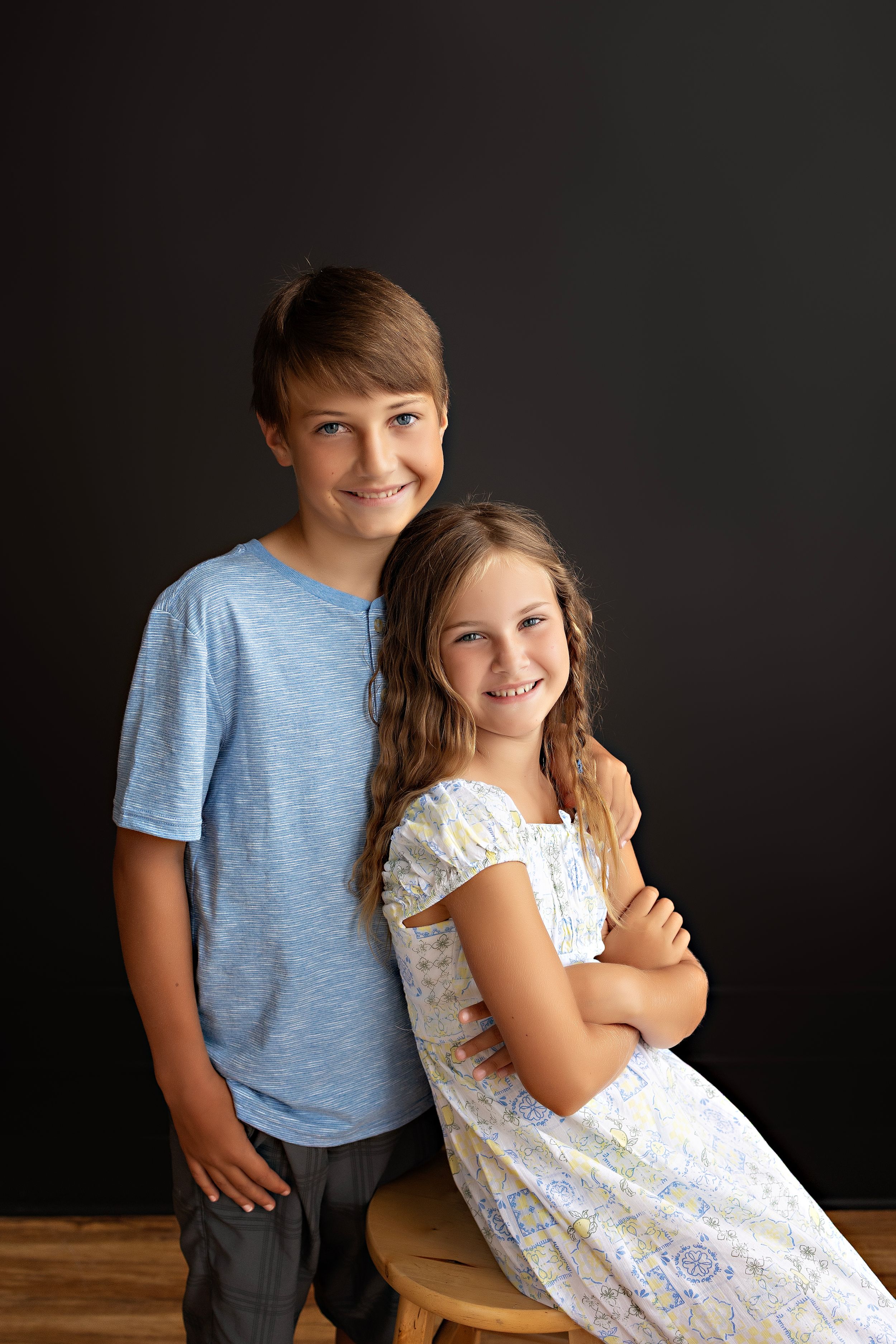 Two young children sitting together in a studio, wearing a blue t-shirt and white lace dress against a dark background.