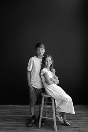 A dramatic black and white sibling photo on a rustic wooden stool against a dark backdrop.