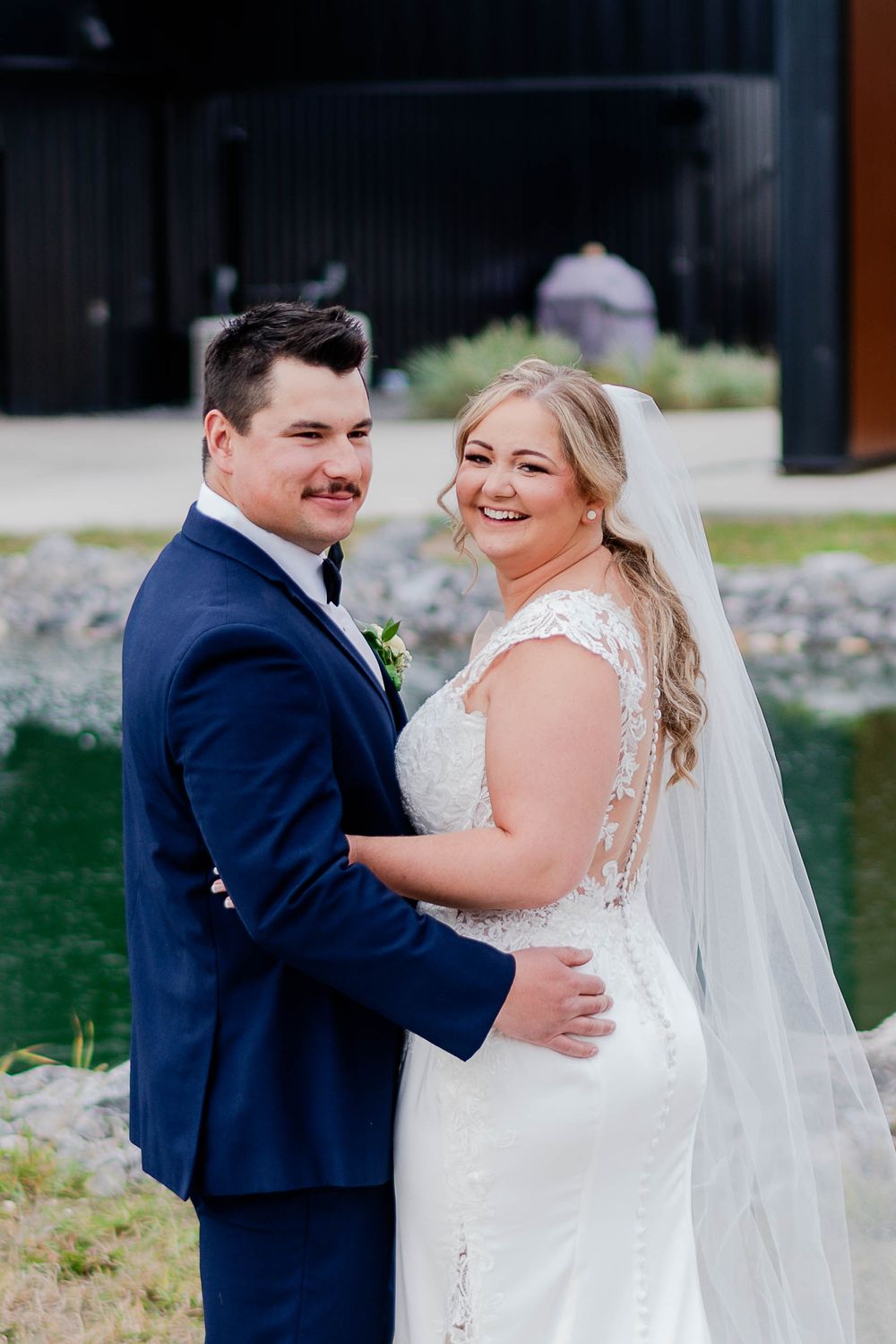A joyful wedding couple embraces outside a modern black building wearing classic formal attire.