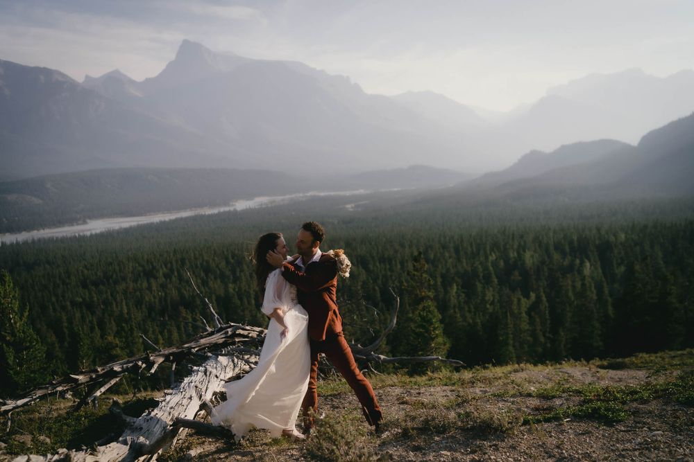 A couple poses dramatically on a mountain overlook with sweeping views of forests and peaks in the background.